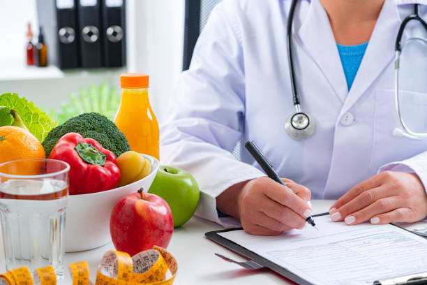 Right nutrition and diet concept. Nutritionist sitting at her desk writing prescription and explaining the benefits of eating fruits and vegetables. An orange juice bottle, glass of water, tape measure are on the doctor's desk and complete the composition. Copy space available for text and/or logo. High resolution 42Mp studio digital capture taken with Sony A7rII and Sony FE 90mm f2.8 macro G OSS lens